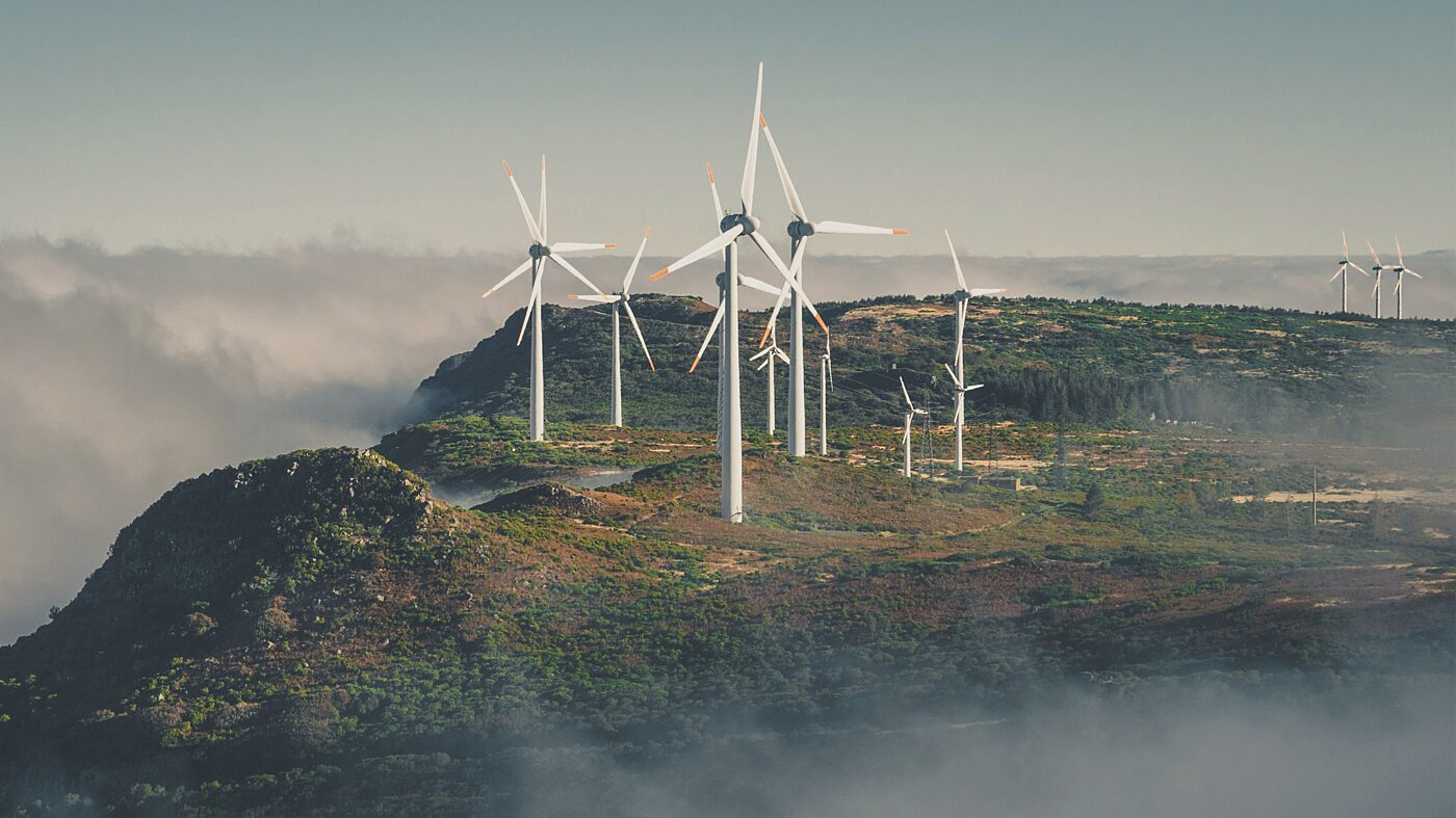A small wind farm perched on top a lush mountaintop; the mountain is surrounded by pillowy fog.